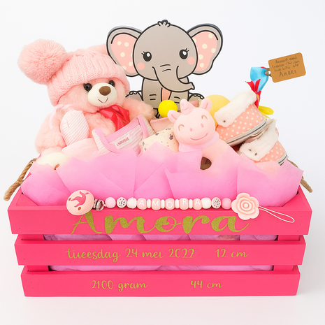 Een handgeschilderde roze houten kist met gouden opschrift “Amora”, gevuld met babycadeaus zoals een beertje, kleertjes, Zw