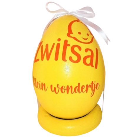 Een felgeel, ei-vormig Zwitsal geschenk met oranje opdruk “Klein wondertje” en wit lint bovenaan, geplaatst op een bijpasse